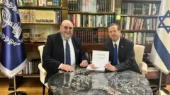 Dois homens sentados à mesa; um deles mostra um documento. Ao fundo, há prateleiras com livros e uma bandeira de Israel