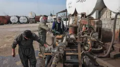 Trabalhadores de um mercado de combustível para automóveis trabalham em um veículo industrial ao lado de tanques de petróleo, em dia de céu azul, em Erbil, no Iraque, no dia 17 de março de 2026