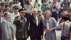 Bivši predsednik SAD Džimi Karter, u crnom odelu sa sivom kravatom i u beloj košulji, sa suprugom Rozalin u sivom kompletu i beloj majici prelazi granicu i ulazi u Severnu Koreju u junu 1994. godine. Oko njih je grupa vojnika i novinari sa fotoaparatima