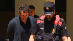 Мужчина в черной кофте с руками за спиной идет, понурив голову, рядом с полицейским в бронежилете.