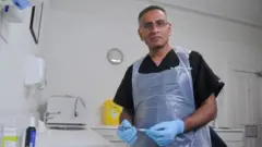 Dalvi Humzah in his white clinical therapy hall, holding a syringe and wearing plastic apron and rubber gloves. In the background, a sink, disorders bucket and disinfectant distributors - the type of healthy resources that are standard in a clinical environment.