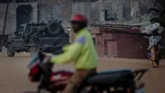 Imodoka itwaye inyeshyamba za M23 ziri ku irondo mu mujyi wa Uvira, ku itariki ya 13 Ukuboza (12) mu mwaka wa 2025. Inyuma yazo ku ruhande hari umugabo uri kuri moto wafotowe mu buryo butamugaragaza neza, hirya ye hari umugore utagaragara mu maso ucyenyeye igitege yiteye ikindi, yikoreye umuzigo muto ku mutwe. Inyuma hirya haraboneka inzu. 