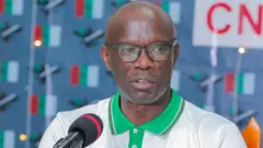 Umukuru w'umugambwe uri ku butegetsi wa CNDD-FDD, Reverien Ndikuriyo yambaye agapira kera, gafise amadebure asa n'urwatsi rubi, yambaye n'amarpori. Imbere yiwe hari mikoro. 