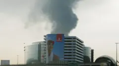 Smoke rising from an area near Dubai International Airport is seen through a vehicle’s windshield after a drone attack hit a fuel tank. In the foreground, an Emirates poster portraying a female flight attendant covers almost the entire façade of a building