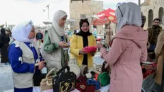 Üç kadın bir tezgahta çantalara bakıyor, pembe paltolu bir kadın onlara ürünleri anlatıyor