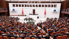 Mecliste oturan insanlar, Cumhurbaşkanı Erdoğan kürsüde konuşuyor.
