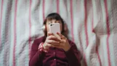 devojčica gleda u telefon dok leži na krevetu