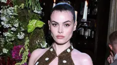A modelo Charli Howard posa com um vestido dourado brilhante com bolinhas brancas e o cabelo castanho penteado para trás em um coque