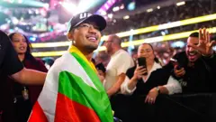 ၂၀၂၅ ခုနှစ်၊ ဒီဇင်ဘာ ၆ ရက်တုန်းက အမေရိကန်ပြည်ထောင်စု နဗားဒါးပြည်နယ်မှာ ကျင်းပတဲ့ ပြိုင်ပွဲအပြီးမှာ အဖြူ အစိမ်း အနီ အဝါရောင် မြန်မာအလံကိုခြုံ UFC တံဆိပ်ပါတဲ့ဦးထုပ်ကို ဆောင်းပြီး ပရိသတ်ဘက်ကို ပြုံးပြနေတဲ့ ဂျော့ရှူဝါ ဗန်၊ သူနောက်မှာ ပရိသတ်တွေက ဖုန်းရဲ့ ရိုက်ပြီး အားပေးနှုတ်ဆက်နေ 