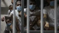 中米エルサルバドルの刑務所に収監されている男性たちを、鉄格子の反対側から写した写真。頭を剃られ、マスクを着け、白いTシャツと短パンを着た人々が、金属製の段ベッドの上に座っている。大勢が腕や脚に入れ墨を入れている