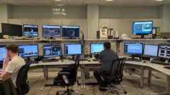 Dos personas observan pantallas de computador en lo que parece ser una sala de control.
