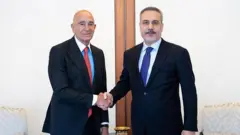 Hakan Fidan ve Amerikalı büyükelçi el sıkışıyor ve kameraya bakıyor.