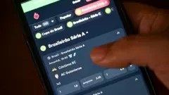 Mão segurando um celular com um aplicativo de apostas na tela, mostrando dados referentes a um jogo de futebol do Campeonato Brasileiro