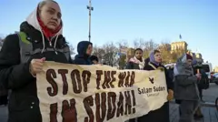 تبدو في الصورة فتاتان ترتديان ملابس شتوية، بشرتهما بيضاء، تحملان لافتة متوسطة الحجم مكتوب عليها باللغة الإنجليزية أوقفوا الحرب في السودان، وحولهما عدد من المتظاهرين. الصورة التقطت نهاراً والسماء زرقاء صافية.