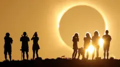 A total solar eclipse is about to occur - a darkened grey sphere appears large in the sky and bright yellow light is emanating from it drenching the sky. Two groups are gathered watching it, silhouetted against the sky 