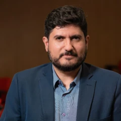 Guilherme Casarões em retrato com semblante sério, camisa de botão e blazer azuis.