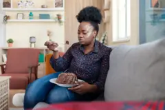 Une jeune femme portant un jean pantalon avec une chemise noire avec des grain orange, tient un plat dans lequel se trouve du chocolat. Elle porte un morceau du chocolat vers la bouche avec une fourchette.