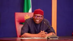 Rivers State Govnor, Siminialayi Fubara wear black cloth with red cap, e sidon for chair dey smile wit im hands crossed. Nigeria flag dey for di background.