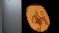 Tüzgiray ailesinin fotoğrafının yer aldığı ahşap bir işleme. Baba Tüzgiray'ın üzerinde takım elbise, anne Tüzgiray'ın üzerinde elbise var. Çocuklar da benzer şekilde şık giyinmiş.