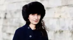 Ayşegül Savaş siyah bir şapka ve paltoyla kameraya bakıyor