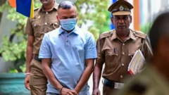 আদালতে নেওয়া হচ্ছে আকাশী রংয়ের পোলোশার্ট পরা তামিম রহমানকে, তার হাতে হাতকড়া ও মুখে মাস্ক। তার সঙ্গে ও পেছনে পুলিশের দুইজন সদস্য। 