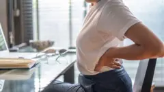 Una mujer sentada en un escritorio se lleva las manos a la espalda en señal de dolor