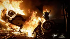 An upturned vehicle blaze sin flames while two silhouetted figures appear to cheer in the background