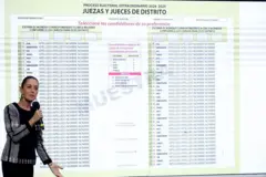 Mulher em pé segurando um microfone. Atrás dela há uma tela que mostra exemplo de uma cédula de votação