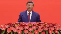 Си Цзиньпин в синем пиджаке, белой рубашке и темно-красном галстуке  во время выступления, перед ним красные цветы и два микрофона. Задний план также красный