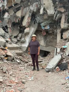 María Lourdes Pérez, vestida con un pantalón y camisa marrones y un gorro beige. Detrás de ella hay una montaña de escombros, hormigón, piedras, partes de una edificación