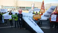 Pessoas protestam segurando cartazes e um jato particular inflável enquanto um ativista usa uma máscara do líder da União Democrata Cristã (CDU) da Alemanha, Friedrich Merz, durante uma manifestação