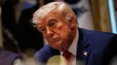 O presidente dos Estados Unidos, Donald Trump, sentado à mesa durante uma reunião, usando terno azul-marinho, camisa branca e gravata vermelha. Ele aparece inclinado para frente, com expressão séria e olhar direcionado para alguém fora do enquadramento.