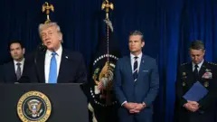 Donald Trump se encuentra en el podio dirigiéndose al público, con una corbata azul y gesticulando con una mano. Detrás de él se encuentran el Secretario de Estado Marco Rubio, el Secretario de Defensa Pete Hegseth y el Presidente del Estado Mayor Conjunto de Estados Unidos, el General Dan Caine, vestido con uniforme militar.