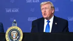 ダークスーツに白シャツ、青色ネクタイを着けたトランプ氏は、米大統領のシールがつけられた演台に立って口を開いている