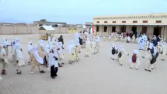 طلاب يرتدون الملابس الرسمية ويحملون حقائبهم، ويشكّلون مجموعات في ساحة المدرسة مع انطلاق الدراسة فيها، وتقف بينهم معلماتهم.