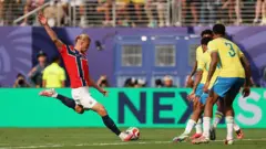 A imagem mostra o momento em que Erling Haaland finaliza de pé esquerdo dentro da área, cercado por quatro jogadores da seleção brasileira. O atacante da Noruega, de uniforme vermelho, aparece no instante do chute, enquanto os defensores brasileiros tentam fechar o espaço à sua frente. Ao fundo, vê-se parte das arquibancadas e os painéis de publicidade ao redor do gramado.