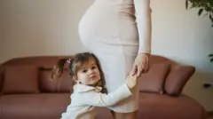 Küçük bir kız çocuğu sadece karnı görünen hamile annesinin ellerinden tutarak ona sarılmış halde ayakta durmuş kameraya bakıyor