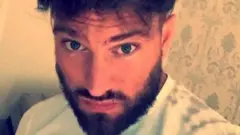 Christopher Harkins olhando para a câmera. Ele tem olhos claros, barba e é um homem branco