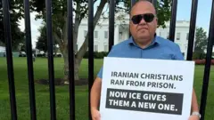 Torosian, con anteojos oscuros, está frente a la Casa Blanca, con una cartel que lee: "Los iraníes cristianos huyeron de prisión; ahora ICE les está dando una nueva".
