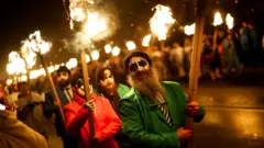 Participants carry fire sticks while wearing masks andf hats and colourful suits during the procession at night.