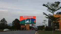 Uma foto ampla do céu, com uma rodovia ao fundo, destaca um painel nas cores laranja e azul com a mensagem “Bem-vindo a Islamabad”