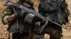 Un homme en tenue militaire tient un fusil. Il porte un insigne arborant le drapeau camerounais.
