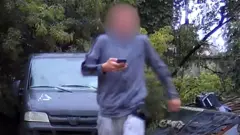 A surveillance photo of a male teenager wearing grey sweat pants and shirt and with his face blurred, carrying a mobile phone. He is walking away from a grey van parked on hard surface in a wooded area.