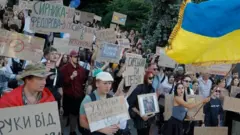 Decenas de personas, en su mayoría jóvenes, portan carteles de cartón en una protesta, mientras una mujer ondea una gran bandera ucraniana.