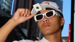 Ảnh chụp cận cảnh một chàng trai trẻ đeo kính trắng và đội mũ lưỡi trai trắng, trên mũ có gắn camera. Anh ta cầm mũ bằng tay phải và nhìn về phía xa, phía sau là những tòa nhà cao tầng.