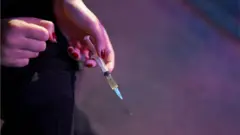 Mão de mulher branca com unhas vermelhas segura uma seringa com um líquido amarelado dentro