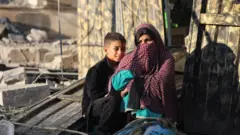إمرأة غزية مع طفلها أمام بيتهما المهدود في مخيم النصيرات