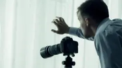 Um homem com uma câmera observa de uma janela.