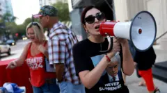 Kobieta o ciemnych włosach i w okularach przeciwsłonecznych, ubrana w czarny T-shirt z wizerunkiem Donalda Trumpa, przemawia przez biało-czerwony megafon. Za nią, w tle po lewej stronie, stoi mężczyzna w białej koszuli w kratę i czapce z daszkiem oraz kobieta o blond włosach ubrana w czerwony top na ramiączkach.