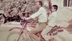 Reza, aged about 11, sitting on the back of a motorcycle with his father driving. He is holding a large gun, pointing it upwards. Both are wearing white shirts. There is a tree, two parked trucks and a building in the background.